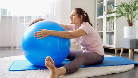 Photo for Beautiful young woman sitting on fitness mat and doing stretching exercises with fitball. Concept of healthcare, sports and yoga at home - Royalty Free Image