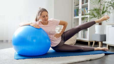 Foto de Mujer joven sonriente sentada en la colchoneta de fitness con el fitball y entrenando sus piernas. Concepto de salud, deportes y yoga en casa - Imagen libre de derechos