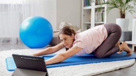 Foto de Mujer joven viendo video en línea entrenamiento deportivo y estiramiento en la estera de fitness. Concepto de salud, deportes y yoga en casa - Imagen libre de derechos