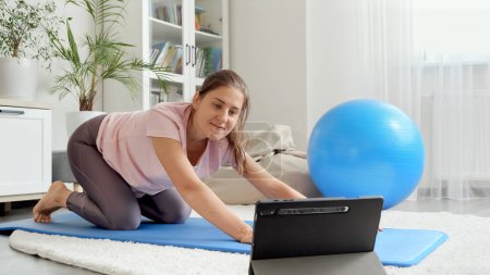Téléchargez les photos : Heureuse femme souriante saluant sa formation sportive en ligne tout en regardant des cours de conditionnement physique sur tablette. Concept de santé, de sport et de yoga à la maison - en image libre de droit