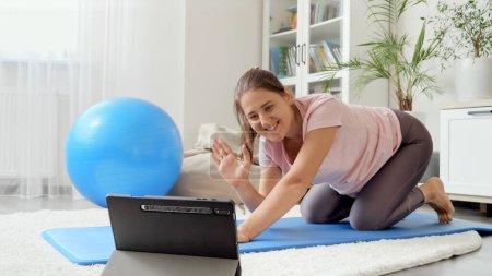Téléchargez les photos : Heureuse femme brune souriante ayant cours de fitness en ligne agitant à la caméra de la tablette. - en image libre de droit