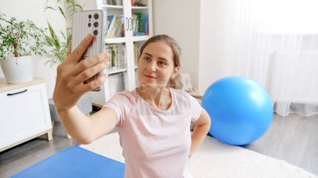 Photo for Young smiling woman makieng selfie photo while doing fitness or yoga at home. - Royalty Free Image