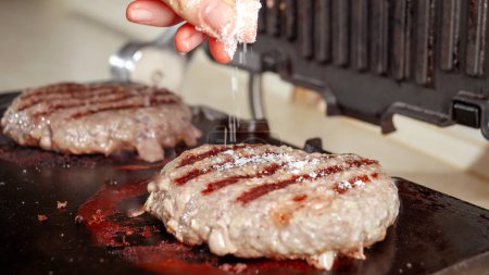 Foto de Primer plano de la mano salazón hamburguesas de carne de res cocinar en la parrilla eléctrica en casa. Cocina en casa, electrodomésticos de cocina, nutrición saludable, ingredientes de hamburguesas - Imagen libre de derechos