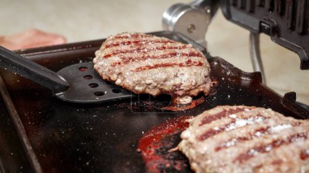 Téléchargez les photos : Gros plan de retourner les galettes de hamburger de bœuf cuire sur le gril électrique. Cuisiner à la maison, appareil de cuisine, alimentation saine, ingrédients pour hamburgers - en image libre de droit