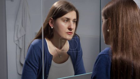 Photo for Sad young brunette woman looking at herself in mirror. Concept of depression, stress, mental illness and problems, loneliness and frustration - Royalty Free Image