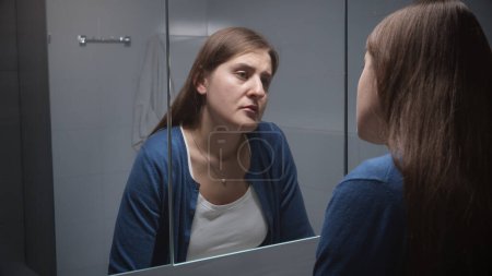 Gestresste Frau in Depressionen lehnt sich an Waschbecken und blickt reflektiert in den Spiegel. Konzept von Depression, Stress, psychischen Erkrankungen und Problemen, Einsamkeit und Frustration