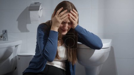 Téléchargez les photos : Jeune femme bouleversée assise dans les toilettes et pleurant. Victime de violence domestique. Dépression féminine - en image libre de droit