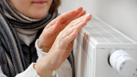 Photo for Closeup of woman freezing at home warming hands at heating radiator. Concept of energy crisis, high bills, broken heating system, economy and saving money on monthly utility payments - Royalty Free Image