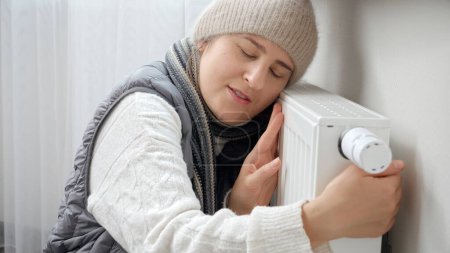 Photo for Young woman in scarf and hat at home embracing heating radiator. Concept of energy crisis, high bills, broken heating system, economy and saving money on monthly utility payments - Royalty Free Image