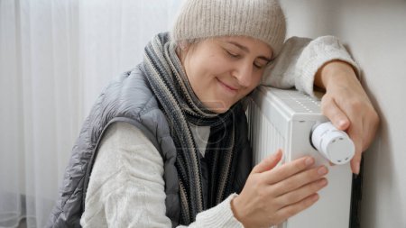 Photo for Young woman feeling very cold at home sitting by the heating radiator. Concept of energy crisis, high bills, broken heating system, economy and saving money on monthly utility payments - Royalty Free Image