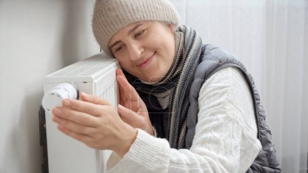 Foto de Retrato de mujer sonriente en ropa de abrigo abrazando radiador de calefacción en casa fría. Concepto de crisis energética, facturas altas, sistema de calefacción roto, economía y ahorro de dinero en pagos mensuales de servicios públicos - Imagen libre de derechos