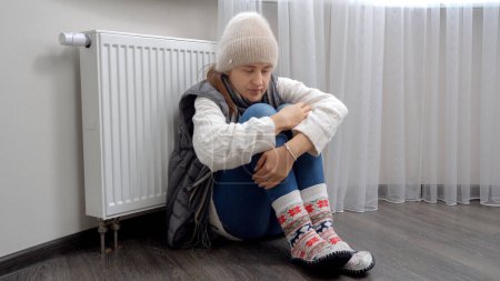 Téléchargez les photos : Jeune femme portant beaucoup de vêtements chauds assis sur le sol à côté du chauffage dans un appartement froid. - en image libre de droit