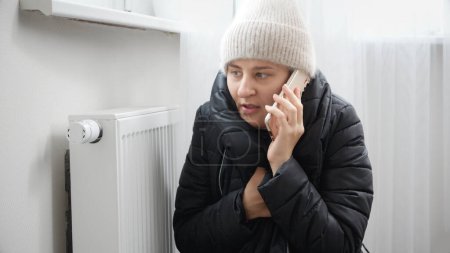 Photo for Portrait of stressed brunette woman in winter coat and woolen hat calling service to repair broken heater radiator. - Royalty Free Image