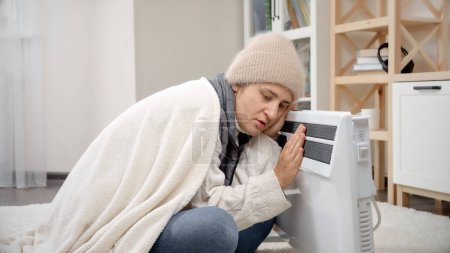 Foto de Mujer joven en ropa de invierno sentada en casa y calentándose en el calentador eléctrico. - Imagen libre de derechos