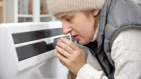 Foto de Retrato de mujer estresada en sombrero caliente sintiéndose fría en casa sentada en el calentador. - Imagen libre de derechos