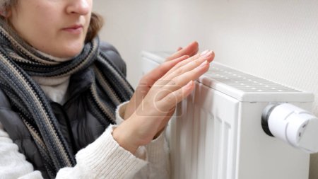 Photo for Closeup of woman in scarf warming hands at heating radiator in house. Concept of energy crisis, high bills, broken heating system, economy and saving money on monthly utility payments. - Royalty Free Image
