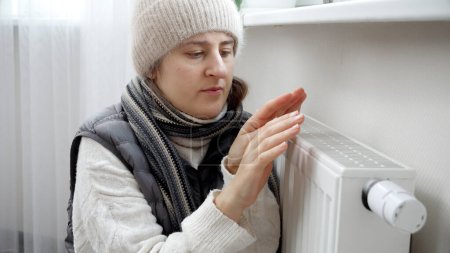 Foto de Retrato de mujer tratando de calentar sus manos frías en radiador de calefacción en piso. Concepto de crisis energética, facturas altas, sistema de calefacción roto, economía y ahorro de dinero en pagos mensuales de servicios públicos - Imagen libre de derechos