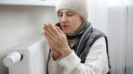 Photo for Young woman in winter clothes and hat feeling cold at home warming at heater or radiator. - Royalty Free Image