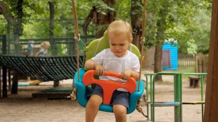 Photo for Portrait of little unhappy boy swinging in colorful swing at park. Kids playing outdoors, children having fun, summer vacation and holiday - Royalty Free Image