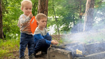 Téléchargez les photos : Deux petits garçons assis au feu de joie dans la forêt et rôtissant du pain sur des bâtons. Loisirs actifs, enfants dans le camping, vacances en famille dans la nature - en image libre de droit