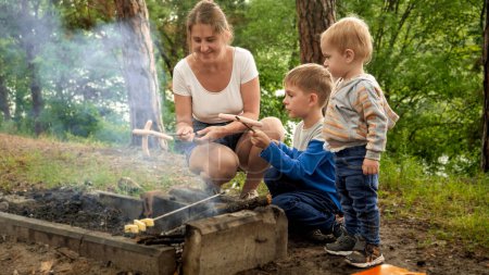 Photo for Happy smiling mother with two boys cooking sausages on the bonfire at forest camp. Active leisure, children in camping, family vacation in nature - Royalty Free Image