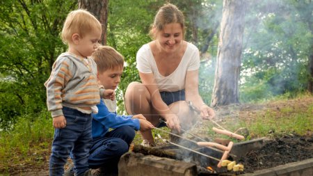 Téléchargez les photos : Deux garçons regardant leur mère cuisiner des saucisses sur le feu de camp dans la forêt. Loisirs actifs, enfants dans le camping, vacances en famille dans la nature - en image libre de droit