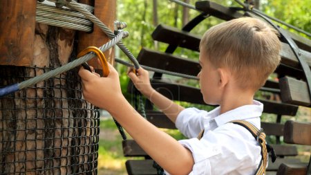 Téléchargez les photos : Portrait de petit garçon serrant son crochet de corde de sécurité tout en grimpant au parc d'escalade extérieur. Enfance active, mode de vie sain, enfants jouant à l'extérieur, enfants dans la nature - en image libre de droit