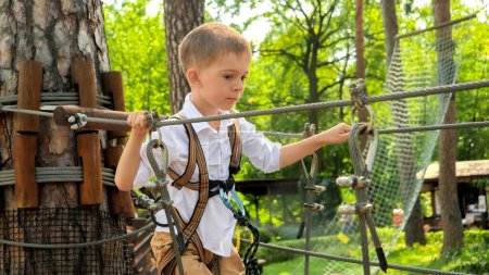 Téléchargez les photos : Portrait de mignon petit garçon marchant sur le pont de corde tout en s'amusant dans le camp scout. Enfance active, mode de vie sain, enfants jouant à l'extérieur, enfants dans la nature - en image libre de droit