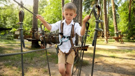 Téléchargez les photos : Petit garçon marchant soigneusement sur le câble ou la corde entre deux arbres dans le parc. Enfance active, mode de vie sain, enfants jouant à l'extérieur, enfants dans la nature - en image libre de droit