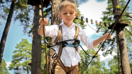 Téléchargez les photos : Portrait d'un petit garçon souriant marchant sur une corde tendue dans un centre d'entraînement d'escalade. Enfance active, mode de vie sain, enfants jouant à l'extérieur, enfants dans la nature - en image libre de droit