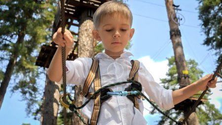 Foto de Niño mirando hacia abajo mientras camina sobre un puente de cuerda de madera en un parque de aventuras extremo. - Imagen libre de derechos