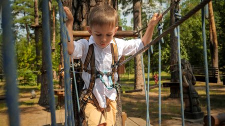 Téléchargez les photos : Petit garçon tenant la corde tout en traversant le pont suspendu entre deux arbres dans le parc. Enfance active, mode de vie sain, enfants jouant à l'extérieur, enfants dans la nature - en image libre de droit