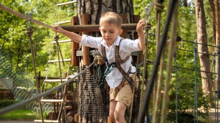 Foto de Feliz chico sonriente divirtiéndose escalando y cruzando obstáculos en el parque de cuerdas. Infancia activa, estilo de vida saludable, niños jugando al aire libre, niños en la naturaleza - Imagen libre de derechos