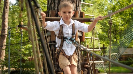 Téléchargez les photos : Portrait d'un petit garçon qui s'amuse en traversant un pont en corde dans un camp d'été. Enfance active, mode de vie sain, enfants jouant à l'extérieur, enfants dans la nature - en image libre de droit
