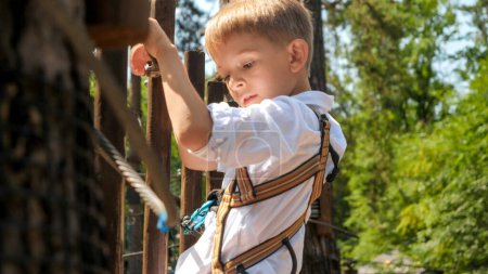 Foto de Retrato de un pequeño oy sosteniendo firmemente la cuerda mientras acaricia los obstáculos en el parque de aventuras. - Imagen libre de derechos