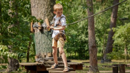 Foto de Niño sosteniendo una cuerda de seguridad y caminando sobre un puente de madera tambaleante en el parque de aventuras. Infancia activa, estilo de vida saludable, niños jugando al aire libre, niños en la naturaleza - Imagen libre de derechos