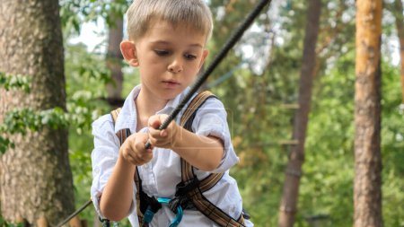 Foto de Retrato de un niño sosteniendo una cuerda apretada mientras cruza obstáculos en el parque de aventuras al aire libre. Infancia activa, estilo de vida saludable, niños jugando al aire libre, niños en la naturaleza - Imagen libre de derechos