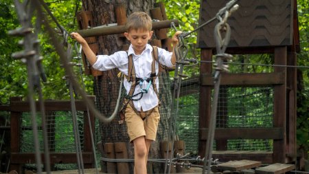 Foto de Lindo chico que se divierte mientras camina en el puente de cuerda oscilante en el parque de aventuras. Infancia activa, estilo de vida saludable, niños jugando al aire libre, niños en la naturaleza - Imagen libre de derechos