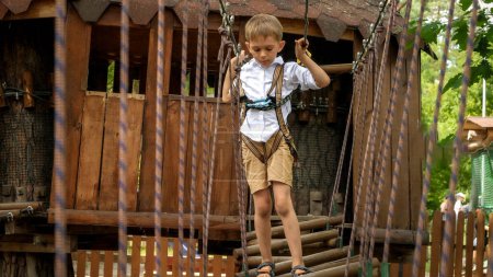 Foto de Un niño sosteniendo una cuerda de seguridad y cruzando un puente tambaleante en el campamento de verano. Deportes para niños, vacaciones de verano, diversión al aire libre, exploradores - Imagen libre de derechos