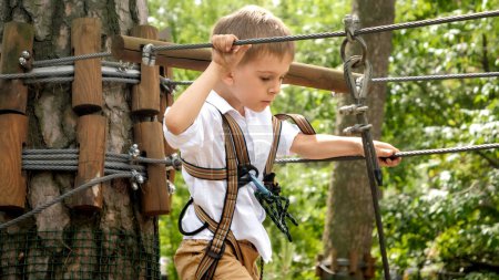 Téléchargez les photos : Portrait d'un garçon courageux marchant sur des obstacles dans un parc à cordes en forêt. Sports pour enfants, vacances d'été, plaisir en plein air, dépisteurs - en image libre de droit