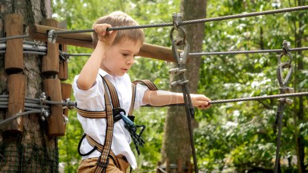 Photo for Little boy hooking safety rope before passing obstacles at rope adventure park. Kids sports, summer holiday, fun outdoors, scouts - Royalty Free Image