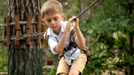 Photo for Portrait of little boy holding tight safety cable while walking on rope bridge at extreme adventure park. Kids sports, summer holiday, fun outdoors, scouts - Royalty Free Image