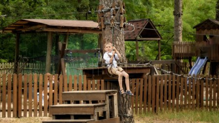 Foto de Retrato de niño alegre feliz cabalgando en la tirolina en el parque de aventura extrema. Deportes para niños, vacaciones de verano, diversión al aire libre, exploradores - Imagen libre de derechos