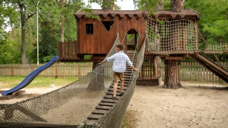 Foto de Feliz niño sonriente caminando en el puente colgante de madera en el parque infantil. Deportes para niños, vacaciones de verano, diversión al aire libre, exploradores - Imagen libre de derechos