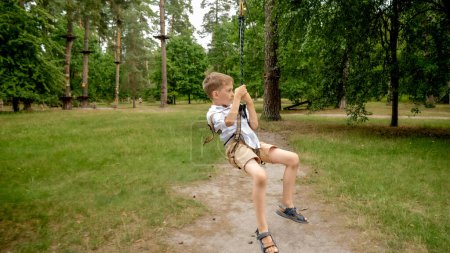 Photo for Cheerful laughing boy riding on the zip-line strung between two trees in adventure park. Kids sports, summer holiday, fun outdoors, scouts - Royalty Free Image