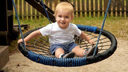 Cute smiling baby boy swinging in rope swing. Kids playing outdoors, children having fun, summer vacation.