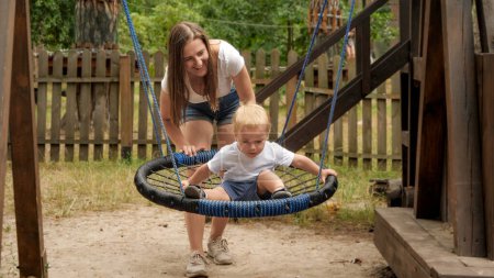 Téléchargez les photos : Rire mère poussant son fils tout-petit balançant sur l'aire de jeux au parc. - en image libre de droit