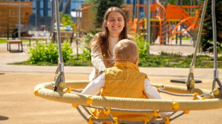 Foto de Joven mujer sonriente feliz expulsando a su hijo pequeño balanceándose en el patio de recreo. - Imagen libre de derechos