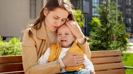 Téléchargez les photos : Jeune mère consolant et caressant son bébé en pleurs sur le banc du parc. Enfants bouleversés, émotions négatives, problèmes d'enfants - en image libre de droit