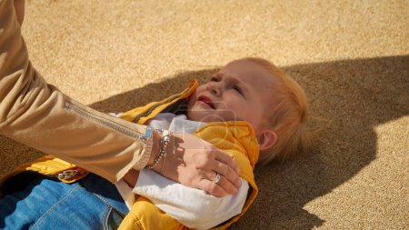 Foto de Joven madre acariciando y consolando a su hijo bebé llorando y gritando en el suelo al aire libre. Niños molestos, emociones negativas, problemas con los niños - Imagen libre de derechos